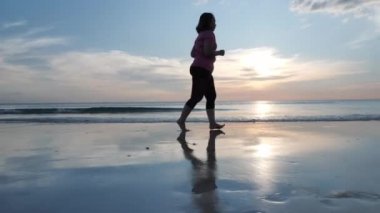 Sahilde gün batımında çıplak ayaklı, çıplak ayaklı Asyalı kadınlar koşuyor. Sağlık egzersizi Yavaş çekim