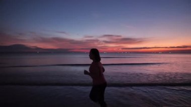 Yandan bakıldığında, çıplak ayaklı, spor elbiseli, şişman Asyalı kadınlar plajda koşuyor. Akşam güneşinde, alacakaranlıkta. Sağlık egzersizi Yavaş çekim