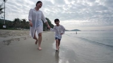 Yaz tatilindeki mutlu Asyalı aile anne ve kızı sabah gün doğumunda kumsalda el ele yürüyorlar. Tatil ve seyahat konsepti. Yavaş çekim