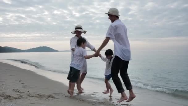 Happy Asian Family On Summer Vacation Father, mother, daughter and son Holding hands jump Spinning around together on the beach In the morning time, sunrise. Holiday and Travel concept. Slow motion 