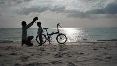Yaz Tatili 'nde Asyalı bir aile plajda bisiklet sürerken oğlu için şapka takıyor. Tatil ve seyahat konsepti. Siluet Yavaş Hareketi