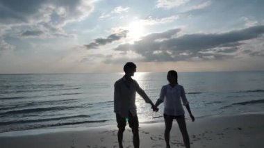 Mutlu Asyalı genç çift el ele tutuşuyor ve birlikte göz teması kuruyorlar. Sahilde yürüyorlar. Denizde günbatımı. Tatil ve seyahat konsepti. Yavaş Hareket