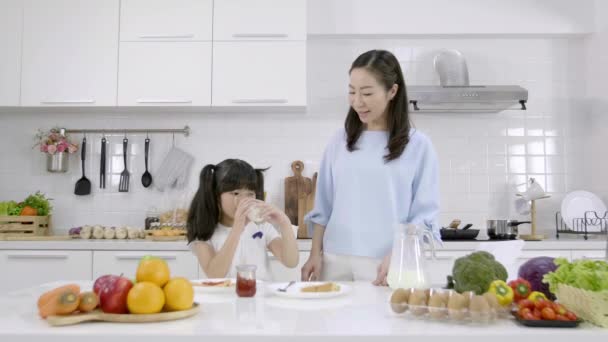 幸せなアジアの家族お母さんと女の子は自宅でキッチンで朝食を食べています パンにジャムと牛乳を飲む 健康食品のコンセプト — ストック動画