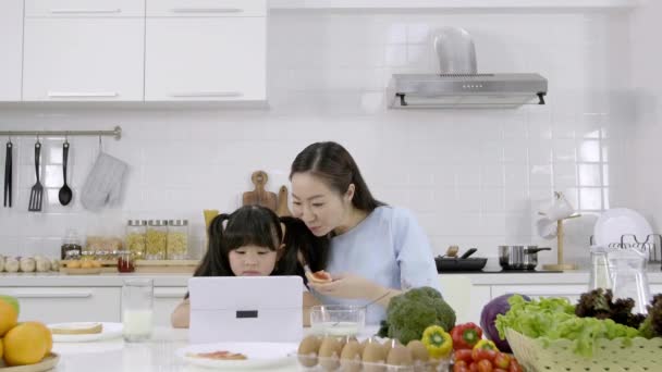 幸せなアジアの家族お母さんと女の子は朝食を食べています 牛乳とシリアル ジャムとパンと家庭で台所でデジタルタブレットを参照してください 健康的な食事と朝の活動 — ストック動画