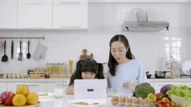 Mutlu Asyalı aile anne ve küçük kız kahvaltı yapıyorlar, mısır gevreği ve süt, reçelli ekmek ve evdeki mutfakta dijital tablet var. Sağlıklı yemek ve sabah aktiviteleri