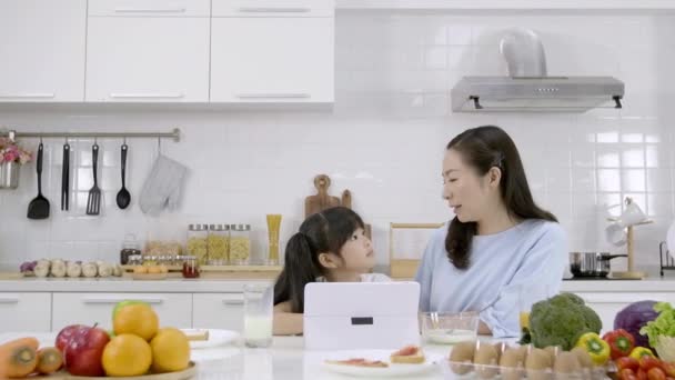 幸せなアジアの家族お母さんと女の子は朝食を食べています 牛乳とシリアル ジャムとパンと家庭で台所でデジタルタブレットを参照してください 健康的な食事と朝の活動 — ストック動画