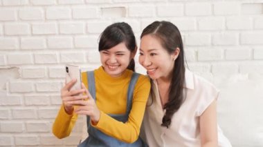 Mutlu Asyalı aile anne ve genç kız akıllı telefonla evde güneş ışığı olan bir odada video konuşuyorlar. İletişim teknolojisi kavramı 