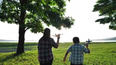 Mutlu Asyalı anne ve oğul, doğada, ağaçlarda ve otlaklarda birlikte uçak simülasyonu yapıyorlar. Akşamları güneş ışığı alan yerlerde. Aile Mutluluğu Tatili Yavaş Hareket