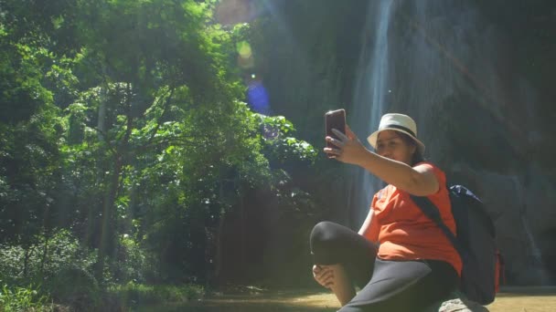 幸せな若いアジアの女性がリラックスし カメラを見て楽しんで 滝でスマートフォンで自撮りやビデオ通話をします 森林旅行の冒険でハイキングするハイカー Asia Thailand — ストック動画
