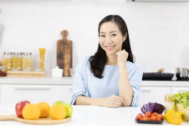 Asyalı bir kadının portresi mutfakta sana gülümsüyor ve evdeki masada salata malzemeleri var. Sağlıklı kadınlar ve sağlıklı gıda konsepti