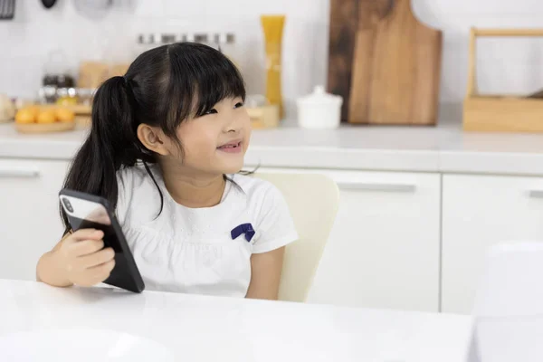 幸せなアジアの女の子は自宅で台所で朝食を準備している母親を待っている間 スマートフォンをお楽しみください テクノロジーと日常生活 ストック写真