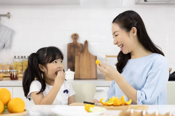 幸せなアジアの家族オレンジフルーツを食べている小さな女の子と母は自宅で台所で野菜や果物を準備しています 健康食品のコンセプト ストック画像