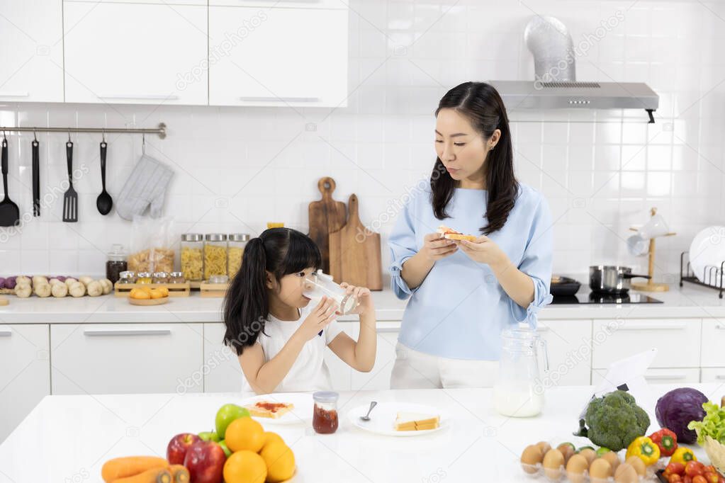 Happy Asian family Mother and Little girl are eating breakfast in the kitchen at home. Bread with jam and drinking milk. Healthy food concept