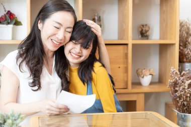 Mutlu Asyalı aile anne ve genç kız kucaklaşır ve evde güneş ışığı olan bir odada kart okurlar. Sevgi gösterin, anneler gününüz kutlu olsun.