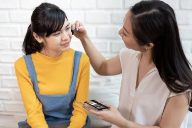 Mutlu Asyalı aile annesi, evde güneş ışığı olan bir odada genç kızına makyaj yapıyor..