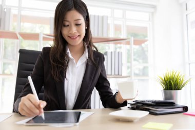 Siyah takım elbiseli Asyalı iş kadınları kahve içerler ve dijital tablete bir mesaj yazarlar. Ofis binasında doğal ışık altında otururlar..