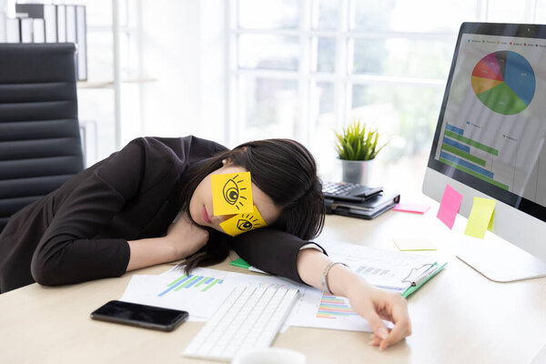 Asian businesswoman in black suit tired frustrated from working at the computer screen. She covering eyes, fake eyes with note pad and Sleeping in desk at office building.