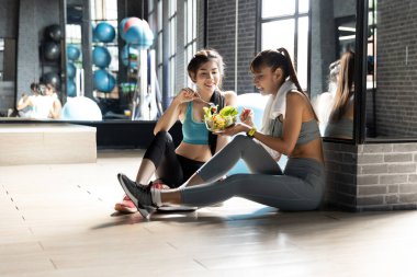 Sağlıklı Asyalı iki genç kadın spor salonunda yeşil salata yiyor penceresi doğal ışık olan bir odada. Fitness ve sağlıklı gıda konsepti