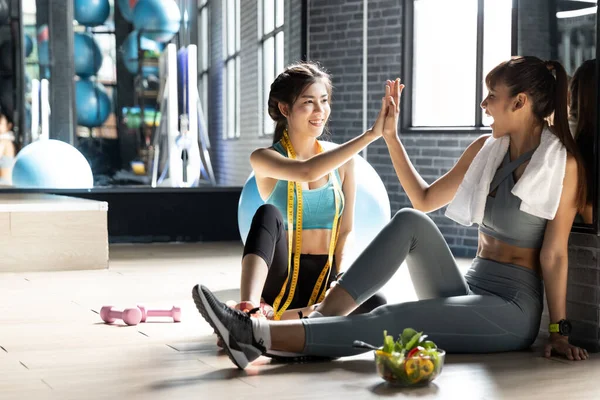 Sağlıklı Asyalı iki genç kadın spor salonundan sonra doğal ışığı olan bir odada beşlik çakıyor. Eğitimden sonra gülümseyen başarılı fitness sınıfı