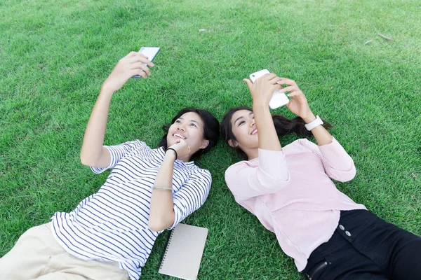 草の上に寝そべっている間に携帯電話のサーフインターネットを使用して幸せな若いアジアの女性2人 ライフスタイル コミュニケーション テクノロジーの概念 コピースペースで ストック写真