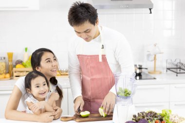 Mutfaktaki mutlu Asyalı aile. Baba, anne ve kız salatayı blender menüsünde lezzetli bir meyve püresi ve evdeki masada malzemeyle hazırlıyorlar. Yemek pişirme, sağlıklı gıda konsepti