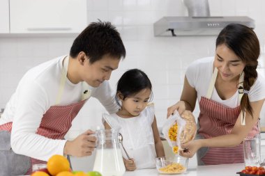 Mutlu Asyalı aile yemek yapıyor. Anne ve baba kahvaltıyı hazırlar, sütle mısır gevreği kızı evde bırakır. Sağlıklı gıda konsepti Vücudun gücü için