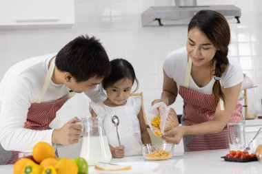 Mutlu Asyalı aile yemek yapıyor. Anne ve baba kahvaltıyı hazırlar, sütle mısır gevreği kızı evde bırakır. Sağlıklı gıda konsepti Vücudun gücü için
