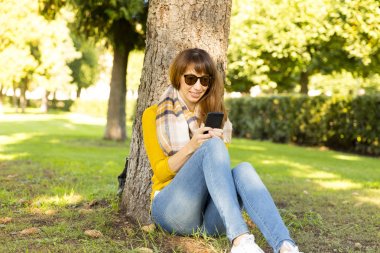 Ağacın yanında oturan ve mesaj yazarak mutlu genç kadın