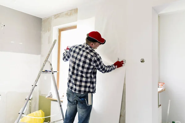 Приклеивание обоев дома. Молодой человек, рабочий вешает обои на стену. Концепция ремонта дома — стоковое фото