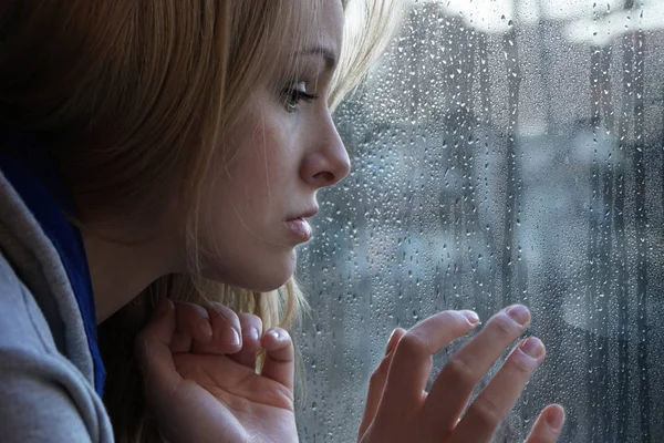 Triste joven mujer mirando a través de la ventana en el día lluvioso —  Fotos de Stock