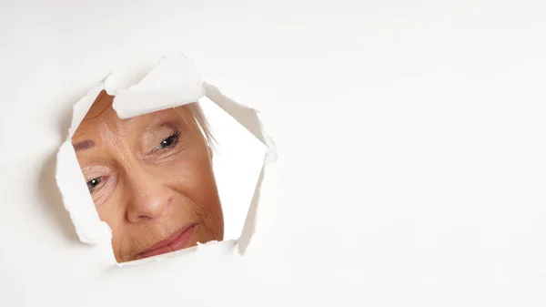 Curious older woman looking through hole at copy space — Stock Photo, Image