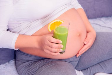 Hamile bir kadın evdeki kanepede dinlenirken sağlıklı organik yeşil meyve suyunun tadını çıkarıyor.. 