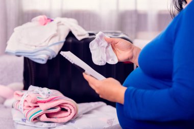 Hamile kadın hastane için yeni doğmuş bebek kıyafetleriyle bavulu toplarken kontrol listesini yapıyor..