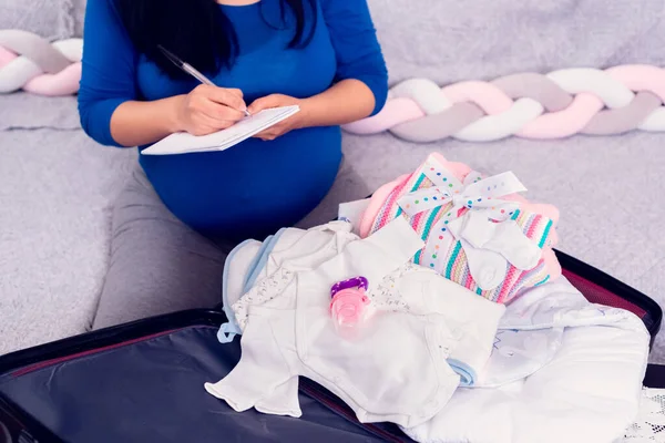 Mujer Embarazada Haciendo Lista Verificación Mientras Empaca Maleta Con Ropa Imagen de archivo