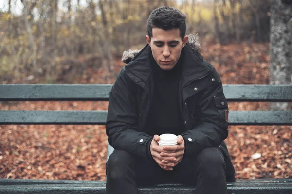 Молодой человек сидел на скамейке с одноразовой чашкой кофе в парке осенью . — стоковое фото