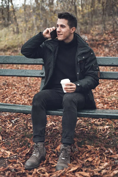 Молодой улыбающийся мужчина в пальто разговаривает по мобильному телефону и пьет кофе на вынос в Осеннем парке . — стоковое фото