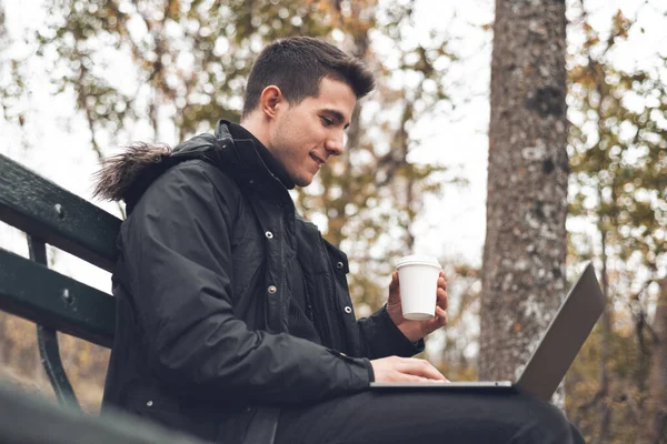 Young man drinking coffee with laptop in autumn park outdoors. Man with laptop and coffee in autumn park under fall foliage. — Stock Photo, Image