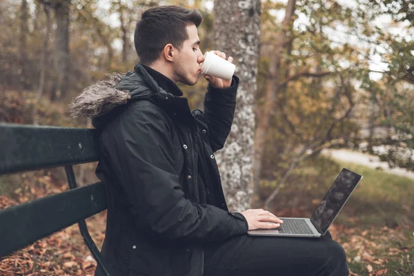 Young man drinking coffee with laptop in autumn park outdoors. Man with laptop and coffee in autumn park under fall foliage. — Stock Photo, Image