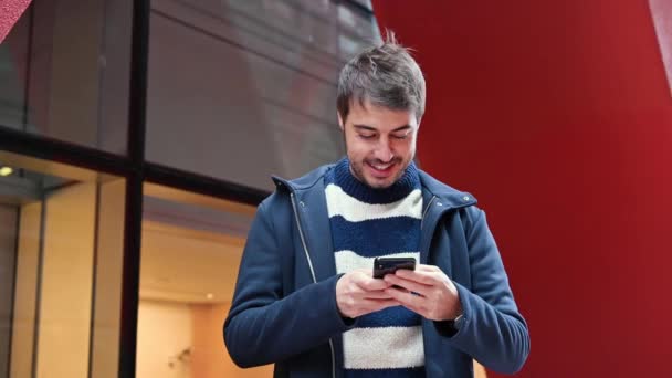 Beau jeune homme, debout devant le bâtiment du bureau, utilisant les réseaux sociaux sur le téléphone portable tout en faisant une pause au travail . — Video