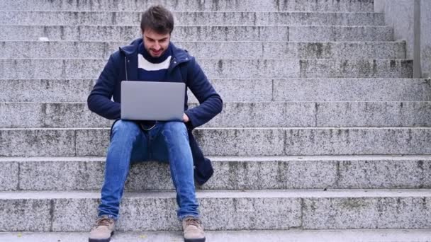 Modny młody człowiek siedzi na kamiennych schodach, pisząc na laptopie.. — Wideo stockowe
