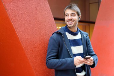 Sokakta duran akıllı telefonu kullanan mutlu bir adam. Arka planda kırmızı renkli modern ofis binaları.