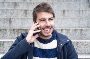 Genç yakışıklı adam, gülümsüyor, telefonda konuşuyor merdivenlerde oturuyor.. 
