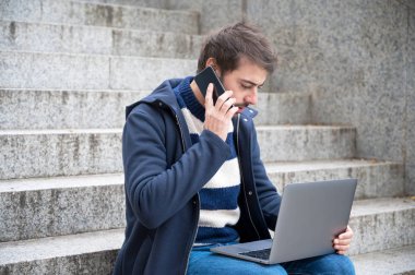 Dizüstü bilgisayarla konuşan adam şehir manzarasında merdivenlerde oturuyor..