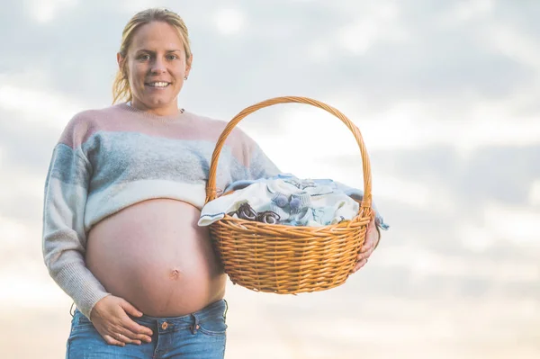 Kobieta w ciąży spodziewa się dziecka, pozowanie na świeżym powietrzu z wiklinowym koszem z ubraniami dla noworodka. — Zdjęcie stockowe