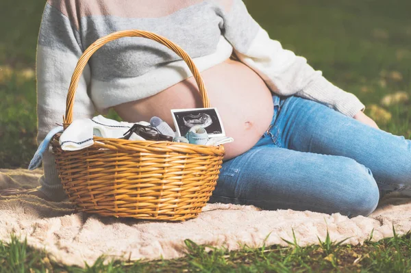Беременная женщина ждет ребенка, позирует на улице с плетеной корзиной с одеждой для новорожденного . — стоковое фото