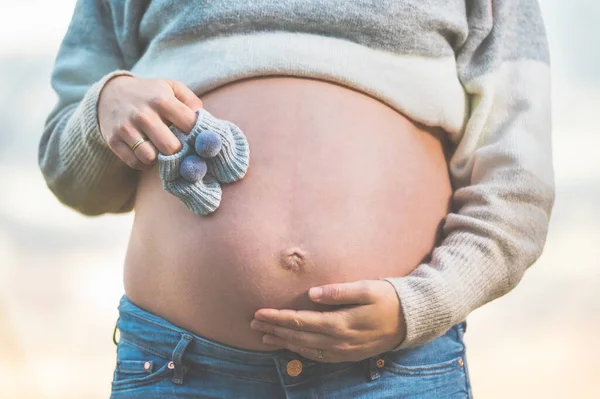 Donna incinta posa all'aperto tenendo piccole scarpe per bambini sulla pancia . — Foto Stock