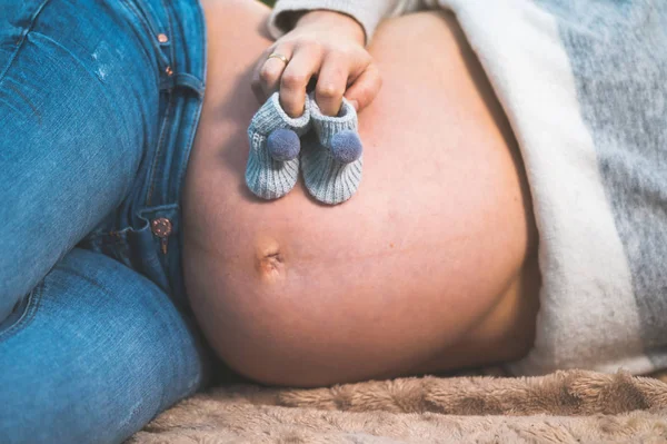 Donna incinta posa all'aperto tenendo piccole scarpe per bambini sulla pancia . — Foto Stock