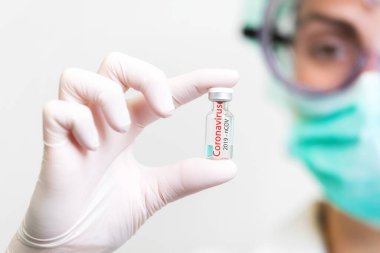 Tıbbi eldivenli ve maskeli bir doktor elinde aşıyla ampula tutuyor. Yakın çekim. Coronavirus, salgın ve tıp konsepti.