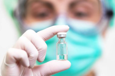 Tıbbi eldivenli ve maskeli bir doktor elinde aşıyla ampula tutuyor. Yakın çekim. Coronavirus, salgın ve tıp konsepti.