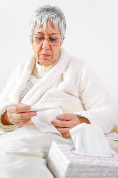 Koronawirus. Chora starsza kobieta trzymająca chusteczkę i termometr siedzi na łóżku, zdenerwowana starsza kobieta złapana na przeziębienie ma objawy grypy koronawirusowej w domu sama. — Zdjęcie stockowe